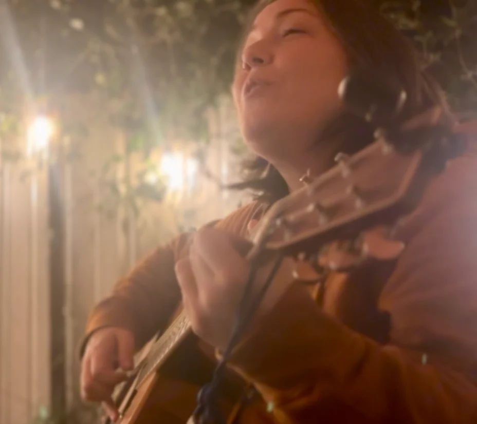 Laura playing acoustic guitar outside at night, eyes closed, singing, surrounded by string lights and jasmine vines on a New Orleans back porch.