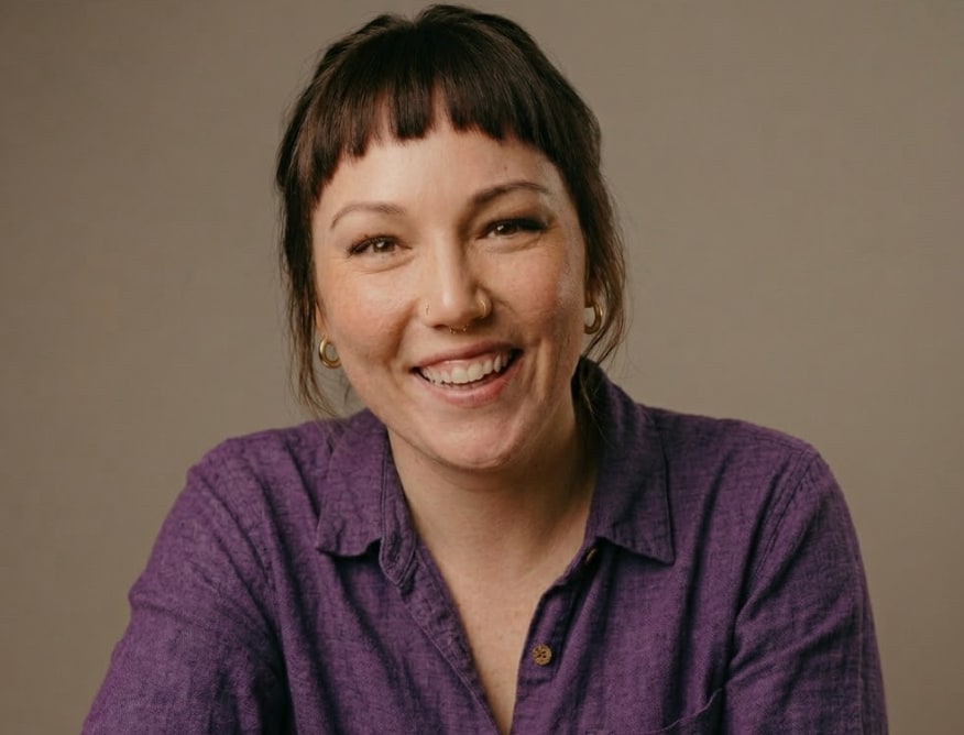 Laura, smiling, in a purple linen shirt against a warm taupe backdrop